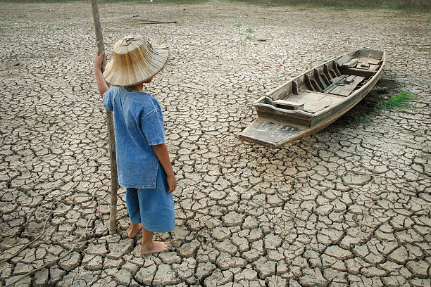 Child in hat on cracked earth near grounded boat, depicting drought impact.